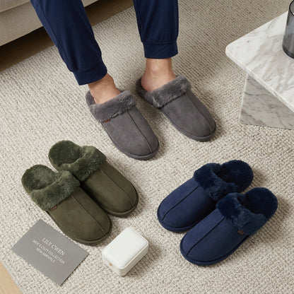 Three pairs of slip-on mule slippers with plush faux-fur trim are arranged on a beige carpet, with a person's lower legs and feet wearing the top pair visible in the scene. The slippers come in gray, olive green, and navy blue.